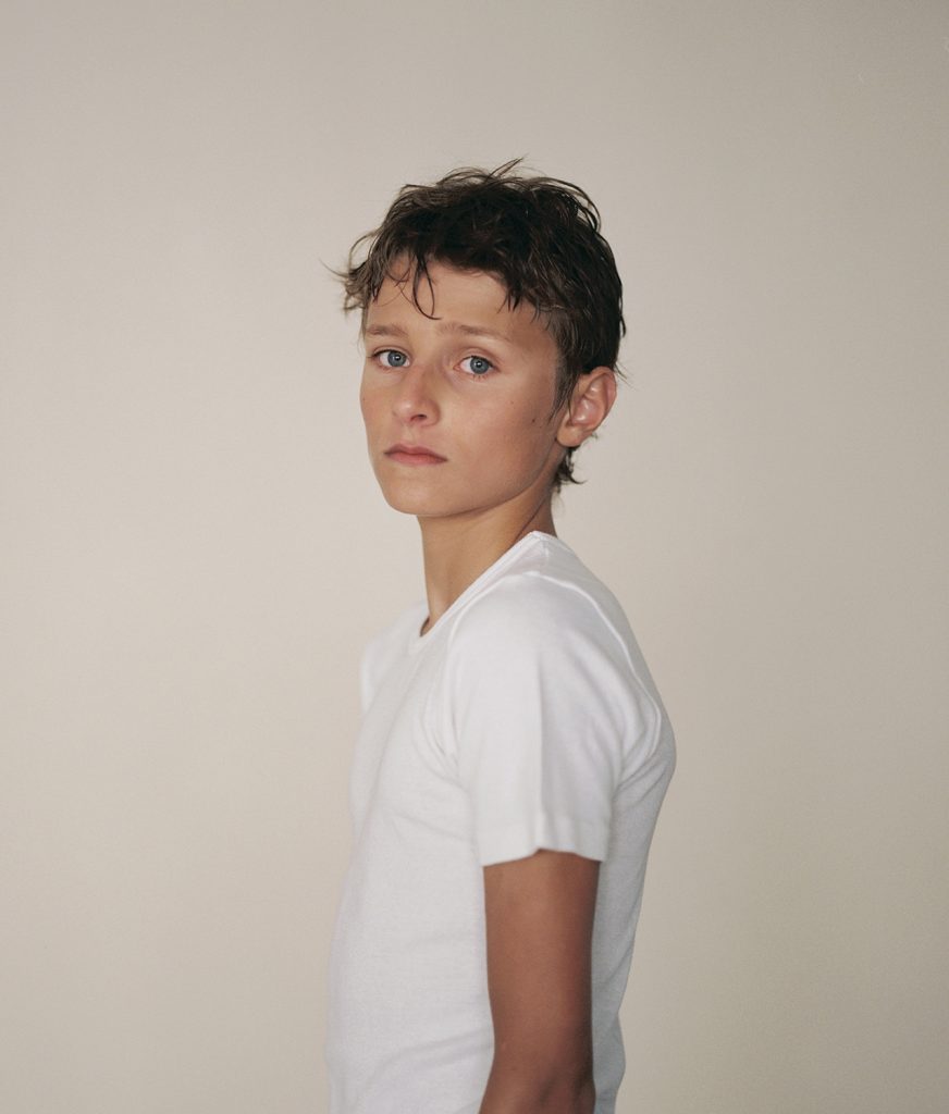 Das Motiv zeigt eine Portraitfotografie aus der Serie "Eliten" von Monika Czosnowska. Zu sehen ist ein Junge mit weißen T-Shirt, der den Betrachter gedankenverloren anschaut. Es handelt sich um einen Schüler einer Eliteschule des Sports aus Berlin.